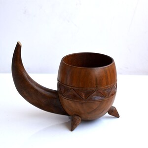 May include: A brown wooden vessel with a rounded bowl, a curved horn-shaped handle extending from one side, and three small pointed feet at the base. The exterior of the bowl features a carved geometric band of triangles around the center.