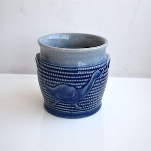 Può includere: Vaso in ceramica blu con un motivo a pois strutturato e un disegno in rilievo di un cigno. Il vaso ha un bordo azzurro e due piccole maniglie. Il cigno è di una tonalità di blu più scura.