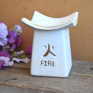 Op de afbeelding: Witte keramische wierookbrander met een gouden rand en het woord "FIRE" erop geschreven. De brander is gevormd als een klein tafeltje met een vierkante basis en een platte bovenkant. Het Chinese karakter voor "vuur" staat ook op de voorkant van de brander.