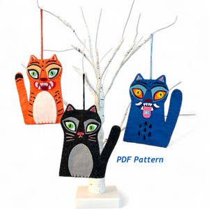 May include: Three felt cat ornaments in orange, black, and blue, each with unique facial expressions and details. The orange cat has tiger stripes, the black cat has green eyes, and the blue cat has a tongue sticking out. The ornaments are hanging from a white tree.