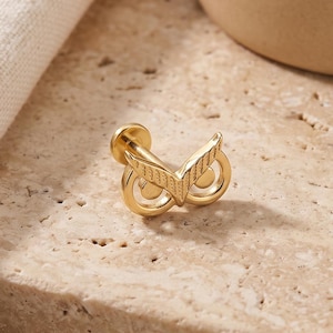 May include: A gold-toned metal body jewellery piece featuring a stylised owl face design with two circular eyes and pointed ears. The jewellery has a flat back post and is designed for piercing, showcasing a polished metallic finish and intricate detailing.