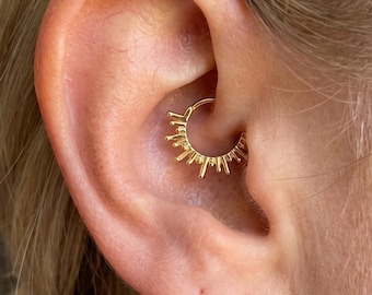 May include: A gold-colored, sunburst-style earring is worn in the ear. The earring is a small hoop with a series of radiating spikes. The ear is against a blurred background.