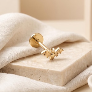 May include: A gold-toned metal flat back stud earring featuring a small bat-shaped charm with outstretched wings. The jewelry piece has a circular base and a straight post, designed for body piercing, showcasing a polished metallic finish.