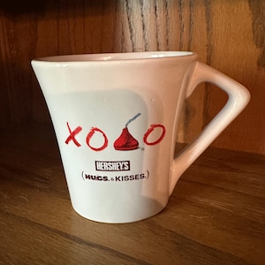 May include: White ceramic mug with a unique handle and a flared shape. The mug features the words "XOXO" in red, a Hershey's Kiss graphic, and the Hershey's logo with the text "(HUGS & KISSES.)".