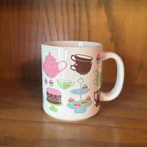 May include: White ceramic mug decorated with pink teapots, brown teacups, green teacups, cupcakes, and a cake. The mug has a white handle and is set against a wooden background. The design is whimsical and colorful.