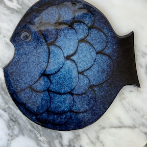 Könnte beinhalten: Ein dekorativer Teller in Form eines blauen Fisches mit schwarzer Schwanzflosse. Der Teller hat ein Schuppenmuster in verschiedenen Blautönen. Er hat ein kleines Loch oben zum Aufhängen.