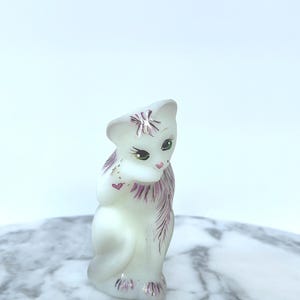 May include: A white ceramic cat figurine with pink and silver accents. The cat has green eyes, pink hearts, and a pink bow on its head. The figurine is sitting, with its paws together, and has pink detailing on its tail and neck.