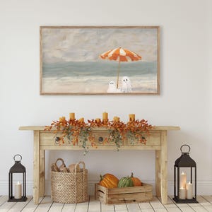 K&ouml;nnte beinhalten: Ein gerahmter Druck zeigt eine Strandszene mit einem orange-wei&szlig; gestreiften Sonnenschirm und zwei Geisterfiguren. Das Kunstwerk h&auml;ngt &uuml;ber einer Holzkonsolentisch, der mit Herbstlaub, Kerzen und K&uuml;rbissen dekoriert ist. Zwei schwarze Laternen mit Kerzen flankieren den Tisch.