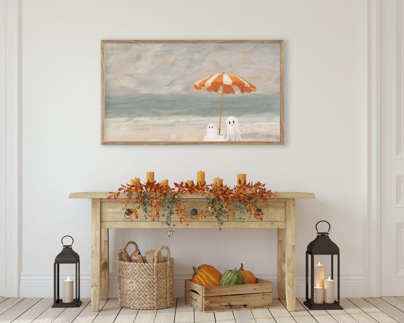 K&ouml;nnte beinhalten: Ein gerahmter Druck zeigt eine Strandszene mit einem orange-wei&szlig; gestreiften Sonnenschirm und zwei Geisterfiguren. Das Kunstwerk h&auml;ngt &uuml;ber einer Holzkonsolentisch, der mit Herbstlaub, Kerzen und K&uuml;rbissen dekoriert ist. Zwei schwarze Laternen mit Kerzen flankieren den Tisch.