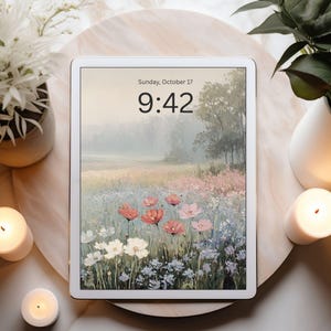 May include: A white tablet displays a landscape painting of a field of wildflowers in shades of pink, white, and blue. The time is 9:42 on Sunday, October 17. The tablet is on a round, light-colored wooden tray, surrounded by candles and plants.