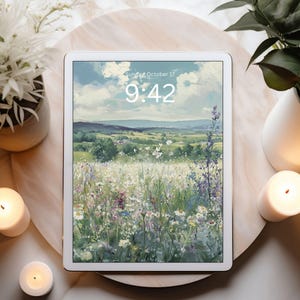 May include: A white tablet displays a landscape painting of a field of wildflowers under a cloudy sky. The time is 9:42 on Sunday, October 17. The tablet is on a marble surface, surrounded by candles and plants.