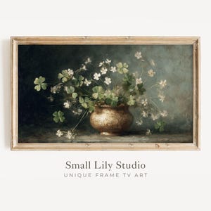 May include: A framed vintage-style print featuring a still life of a bronze-colored vase overflowing with green shamrocks and delicate white flowers. The artwork has a dark, moody background and is labeled "Small Lily Studio".