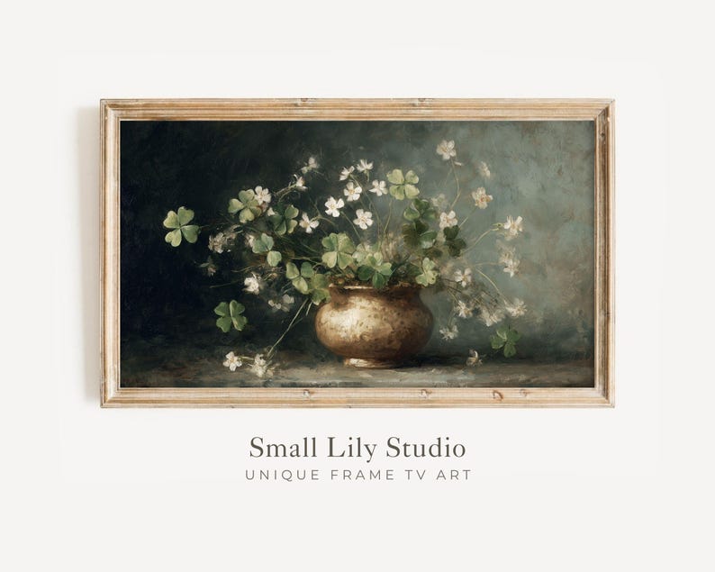 Puede incluir: Una impresi&oacute;n enmarcada de estilo vintage que presenta una naturaleza muerta de un jarr&oacute;n de color bronce rebosante de tr&eacute;boles verdes y delicadas flores blancas. La obra de arte tiene un fondo oscuro y est&aacute; etiquetada como "Small Lily Studio".