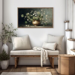 Puede incluir: Una obra de arte enmarcada representa una naturaleza muerta de flores blancas y follaje verde en un jarr&oacute;n dorado. La obra cuelga sobre un banco de madera con cojines y una manta color crema. Tambi&eacute;n se ven una cesta tejida y una escalera.