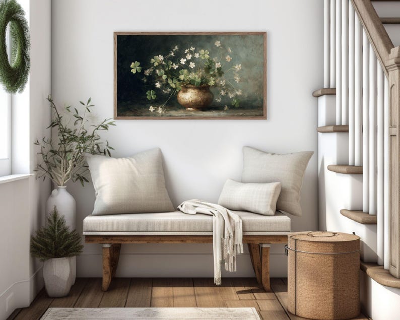 Puede incluir: Una obra de arte enmarcada representa una naturaleza muerta de flores blancas y follaje verde en un jarr&oacute;n dorado. La obra cuelga sobre un banco de madera con cojines y una manta color crema. Tambi&eacute;n se ven una cesta tejida y una escalera.