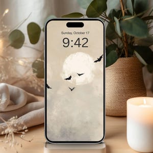 May include: A smartphone displays a Halloween-themed wallpaper featuring a full moon, bats, and a cloudy sky. The time is 9:42 AM on Sunday, October 17. A lit candle and a potted plant are in the background.