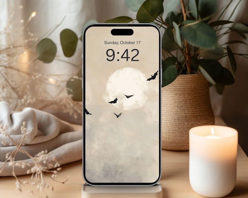 May include: A smartphone displays a Halloween-themed wallpaper featuring a full moon, bats, and a cloudy sky. The time is 9:42 AM on Sunday, October 17. A lit candle and a potted plant are in the background.