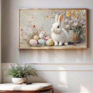 May include: A framed painting shows a white rabbit and a smaller rabbit beside decorated Easter eggs. The artwork uses a soft, pastel colour palette with flowers and foliage, creating a spring theme. The frame is gold.