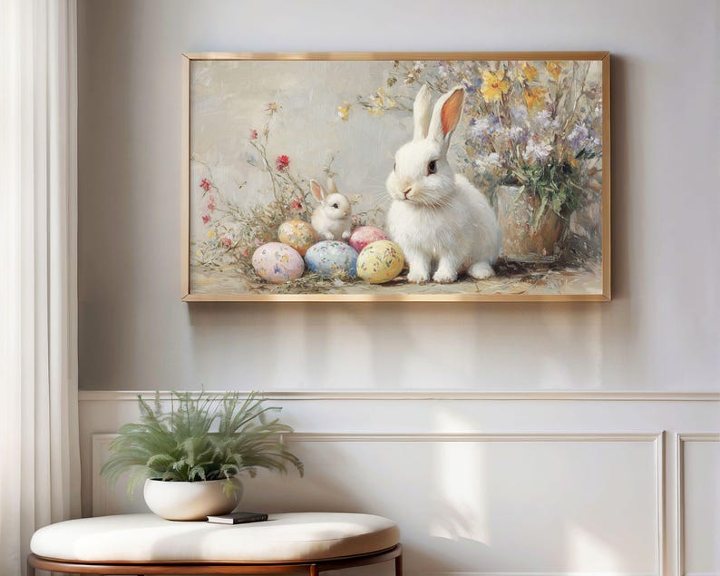 May include: A framed painting shows a white rabbit and a smaller rabbit beside decorated Easter eggs. The artwork uses a soft, pastel colour palette with flowers and foliage, creating a spring theme. The frame is gold.