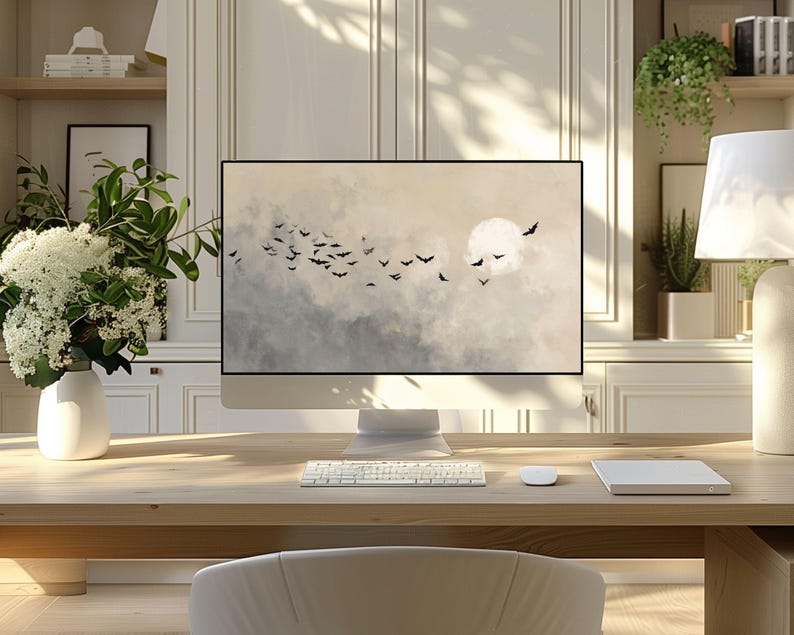 May include: A modern computer monitor displays a serene landscape with a full moon and silhouetted birds against a cloudy sky. The monitor sits on a light wood desk with a keyboard, mouse, and a white notepad. A white vase with flowers is on the left.
