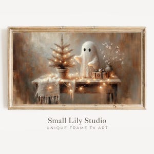 Puede incluir: Una impresión artística digital enmarcada que presenta una escena caprichosa. Un pequeño árbol de Navidad en maceta y una figura de fantasma de peluche están sobre una mesa de madera, adornados con luces de cadena. La obra de arte tiene una paleta de colores cálidos y neutros, con el texto "Small Lily Studio".