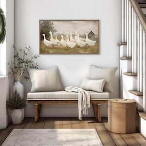 May include: A framed artwork depicts a flock of white geese in a field with a red barn in the background. The artwork hangs above a wooden bench with cream-colored cushions and pillows. A green wreath hangs on the wall near a window.