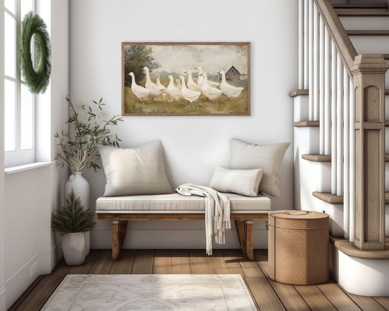 May include: A framed artwork depicts a flock of white geese in a field with a red barn in the background. The artwork hangs above a wooden bench with cream-colored cushions and pillows. A green wreath hangs on the wall near a window.