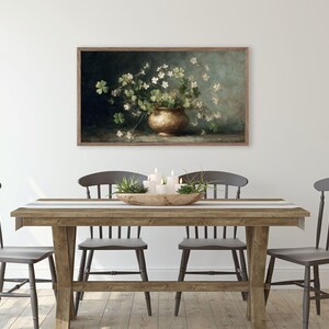 Puede incluir: Una obra de arte enmarcada representa una naturaleza muerta con un jarr&oacute;n de color bronce lleno de flores blancas y tr&eacute;boles verdes. La obra de arte est&aacute; colgada en una pared blanca sobre una mesa de comedor de madera con cuatro sillas grises.