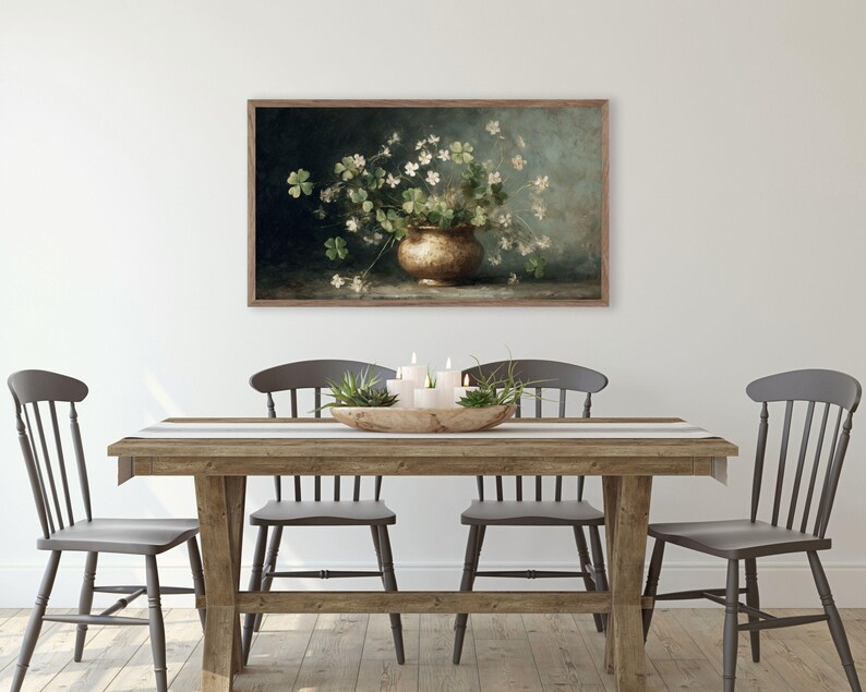 Puede incluir: Una obra de arte enmarcada representa una naturaleza muerta con un jarr&oacute;n de color bronce lleno de flores blancas y tr&eacute;boles verdes. La obra de arte est&aacute; colgada en una pared blanca sobre una mesa de comedor de madera con cuatro sillas grises.