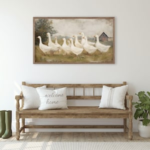 May include: A framed painting depicts a flock of white geese in a field with a red barn in the background. Below, a wooden bench holds striped and text pillows. Green rain boots stand beside the bench.