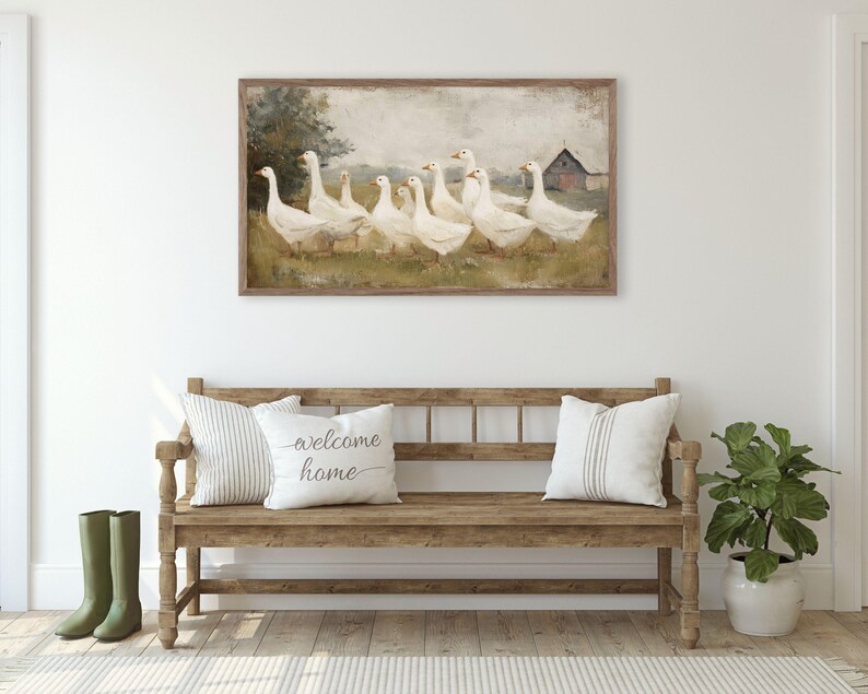 May include: A framed painting depicts a flock of white geese in a field with a red barn in the background. Below, a wooden bench holds striped and text pillows. Green rain boots stand beside the bench.