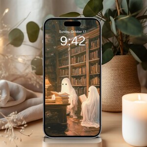 May include: A smartphone displays a spooky library scene with two ghost figures reading books. The screen shows the date and time: Sunday, October 17, 9:42. A lit candle and a potted plant are in the background.