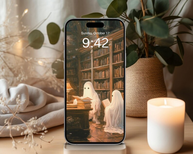 May include: A smartphone displays a spooky library scene with two ghost figures reading books. The screen shows the date and time: Sunday, October 17, 9:42. A lit candle and a potted plant are in the background.