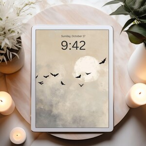 May include: A white tablet displays a digital art scene of a full moon, bats, and clouds. The time is 9:42 on Sunday, October 17. The tablet is surrounded by candles, plants, and a decorative tray, creating a cozy, autumnal atmosphere.