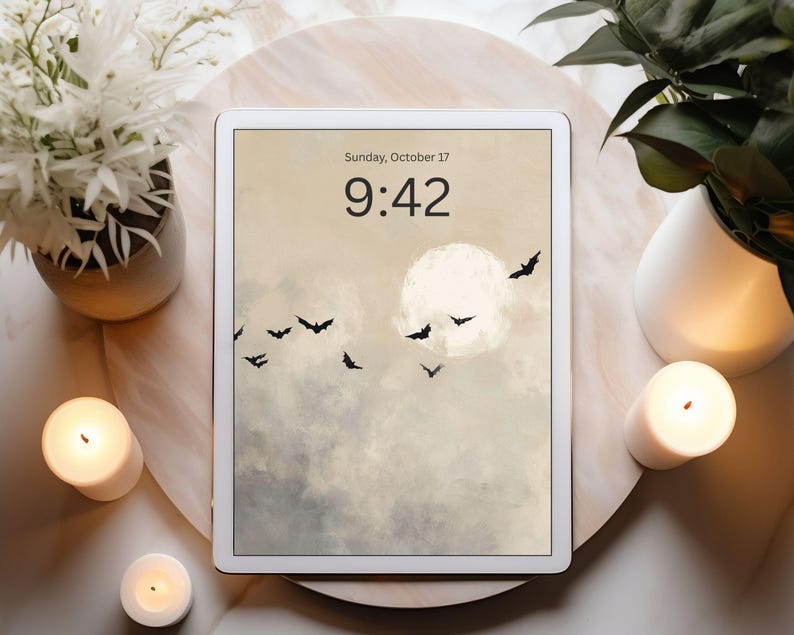May include: A white tablet displays a digital art scene of a full moon, bats, and clouds. The time is 9:42 on Sunday, October 17. The tablet is surrounded by candles, plants, and a decorative tray, creating a cozy, autumnal atmosphere.