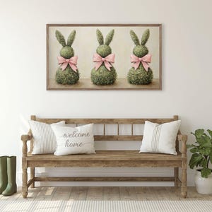 May include: A framed print depicting three topiary rabbit sculptures adorned with pink bows. The artwork is above a wooden bench with cushions, one bearing the text "welcome home". Green wellington boots are placed to the left.