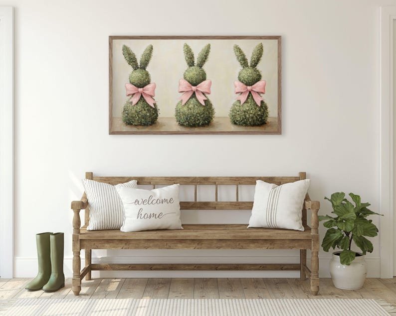 May include: A framed print depicting three topiary rabbit sculptures adorned with pink bows. The artwork is above a wooden bench with cushions, one bearing the text "welcome home". Green wellington boots are placed to the left.