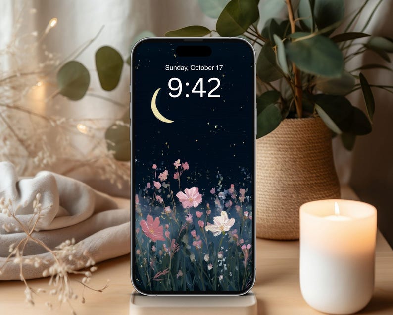 May include: A smartphone displays a floral wallpaper with a night sky, moon, and stars. The time is 9:42 AM on Sunday, October 17. A lit candle and a potted plant are in the background, creating a cozy atmosphere.