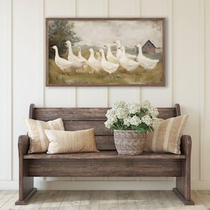 May include: A framed painting depicts a flock of white geese in a field with a red barn in the background. The painting hangs above a wooden bench with pillows and a basket of white flowers.