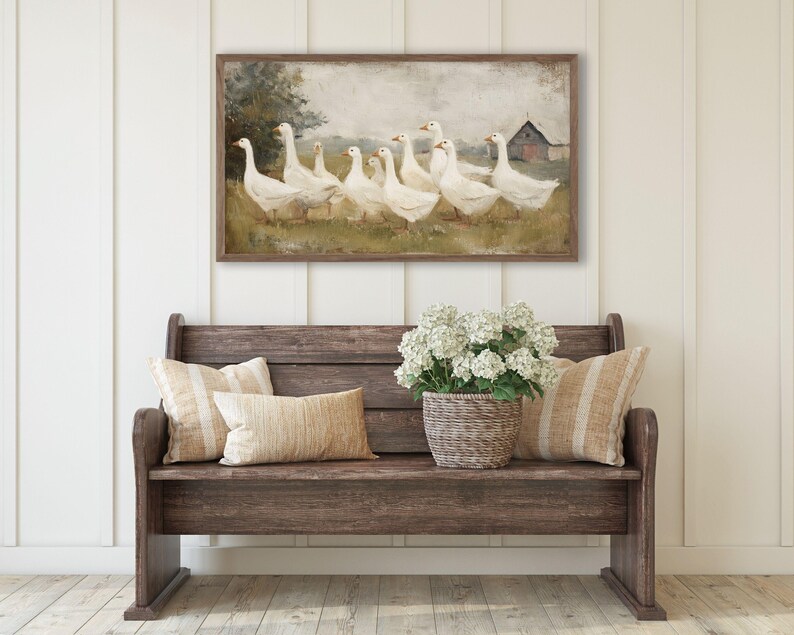 May include: A framed painting depicts a flock of white geese in a field with a red barn in the background. The painting hangs above a wooden bench with pillows and a basket of white flowers.