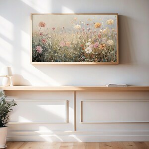 May include: A framed floral print depicting a meadow of wildflowers in pastel hues. The artwork hangs on a white wall above a wooden shelf. The flowers include pink, yellow, and white blossoms.