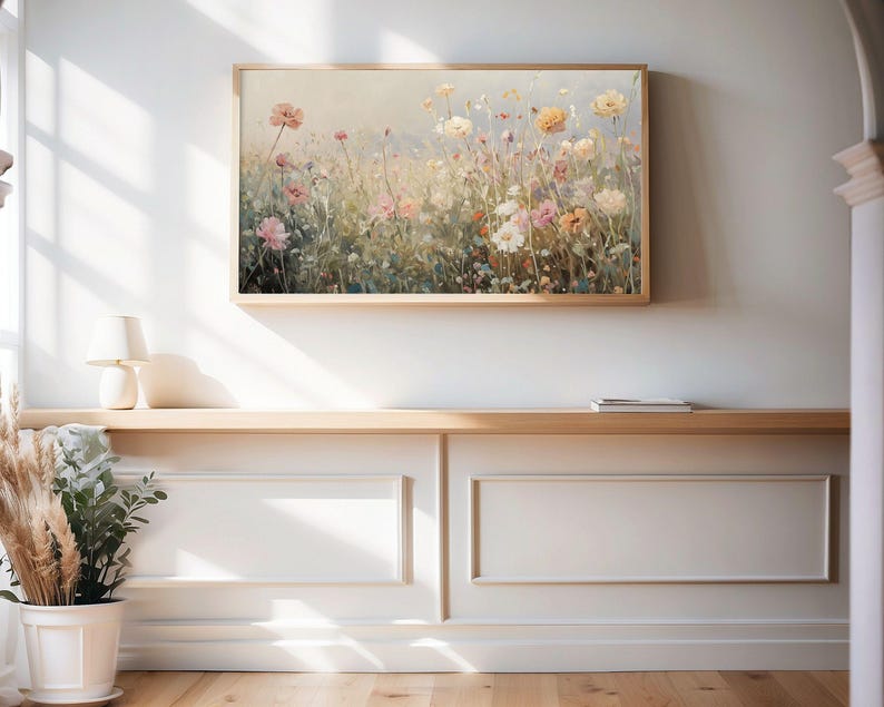 May include: A framed floral print depicting a meadow of wildflowers in pastel hues. The artwork hangs on a white wall above a wooden shelf. The flowers include pink, yellow, and white blossoms.