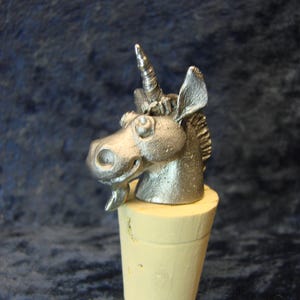 May include: A silver-colored unicorn head bottle stopper. The unicorn has a detailed mane, a spiraled horn, and a smiling expression. The stopper is mounted on a light yellow cork base, perfect for wine bottles.