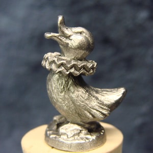 May include: A silver-colored duck figurine with a ruffled collar, mounted on a cork. The duck is depicted with its beak open, as if quacking. The figurine is detailed and stands on a small base.