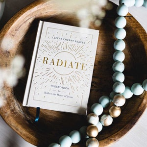 May include: A white book with gold lettering titled "RADIATE 90 Devotions to Reflect the Heart of Jesus" by Cleere Cherry Reaves. The book is lying on a wooden surface with a string of light blue and gold beads.