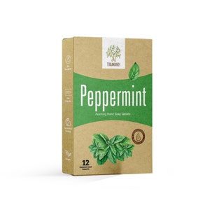 May include: A brown cardboard box with a white label that says "Peppermint Foaming Hand Soap Tablets" and a green leaf design. The box also has a logo for the brand "TERRA MANU" and a small image of a leaf with a drop of water. The box contains 12 tablets.