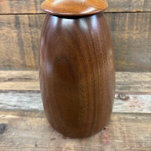Puede incluir: Un recipiente de madera con tapa, hecho de madera de nogal marrón oscuro. La tapa tiene una incrustación de madera marrón claro.