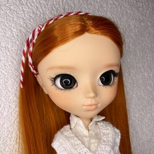 Könnte beinhalten: Puppe mit langem, rotem Haar und großen, ausdrucksstarken Augen. Die Puppe hat einen hellen Teint mit Sommersprossen und trägt eine weiße Strickweste über einem Hemd mit Kragen. Ein rot-weißes Stirnband ziert den Kopf der Puppe.