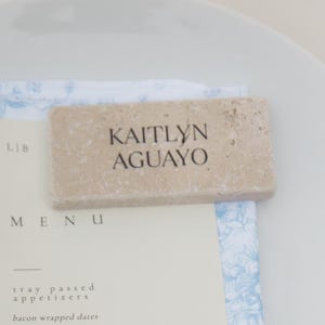 May include: A white plate with a menu and a napkin. The menu features a list of appetizers, buffet items, and desserts. A stone name card with the name "KAITLYN AGUAYO" rests on top. The napkin has a blue floral pattern.