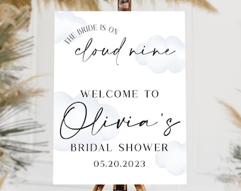 May include: A white sign on a wooden easel with the text "The Bride is on cloud nine. Welcome to Olivia's Bridal Shower 05.20.2023." The sign has watercolor cloud accents. The background includes dried floral arrangements.